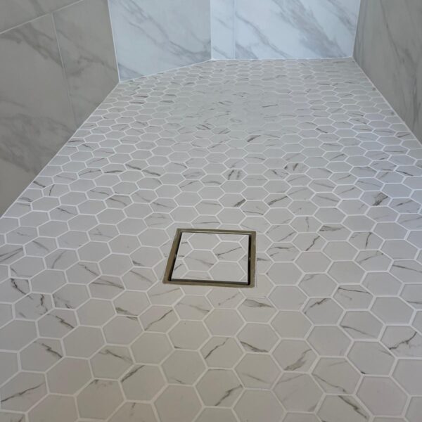 Shower floor with white hexagonal tiles, a square metal drain in the center, and marble-patterned wall tiles—perfect for your bathroom remodel. A small corner bench is visible in the background.