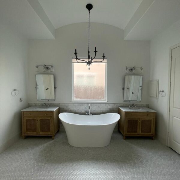 A modern bathroom in The Woodlands features a freestanding white bathtub centered beneath a frosted window, flanked by two wooden vanities with sinks and mirrors, and a black chandelier hanging from the ceiling.