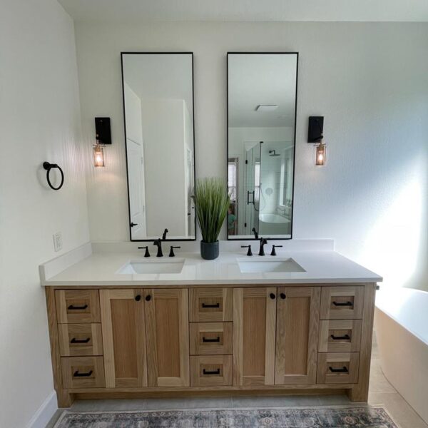 A modern bathroom renovation in The Woodlands featuring a wooden double-sink vanity, two rectangular mirrors, sleek black fixtures, wall sconces on each side, a green plant between the sinks, and a patterned rug on the floor.