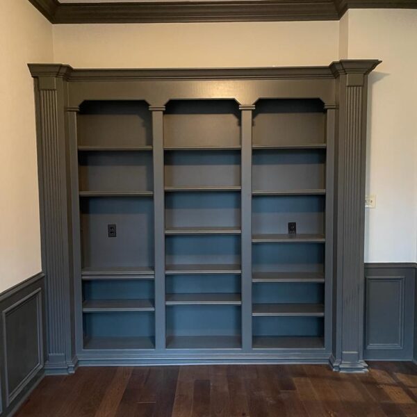 A built-in dark gray wooden bookshelf with three sections, each featuring four shelves, stands against a cream-colored wall with decorative trim, atop a dark hardwood floor—perfect for your next renovation or to complement a modern kitchen space.