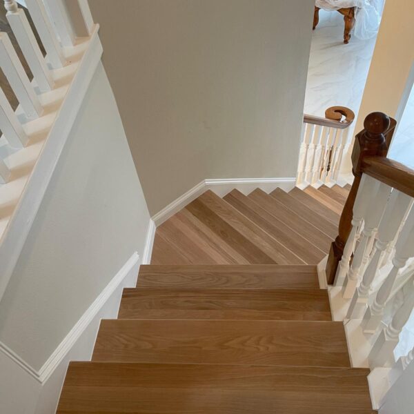 A wooden staircase with white railings turns at a corner, leading down to a brightly lit room with a billiard table covered in a white cloth—a stylish remodel inspired by homes in The Woodlands.