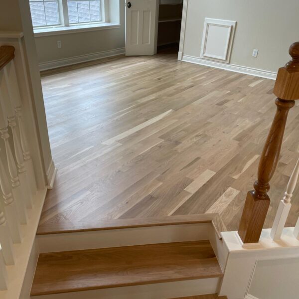 A bright room in The Woodlands with light hardwood floors, a window letting in natural light, white walls, a door to a walk-in closet, and a few stairs with a wooden handrail leading down to the space near the kitchen.