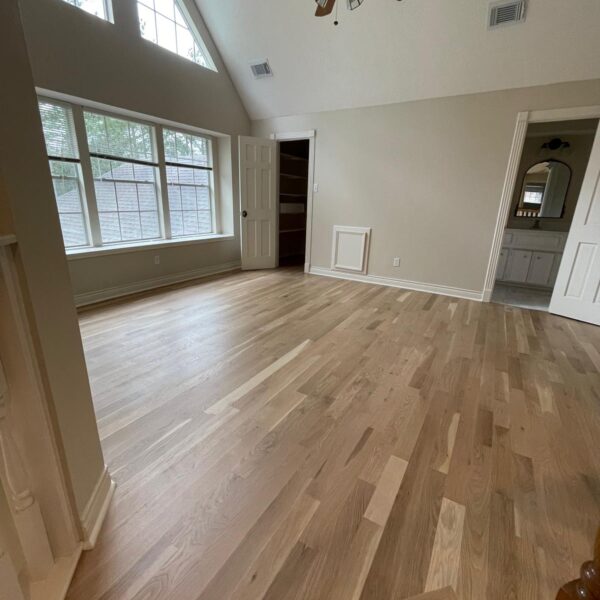 Spacious, empty room with light wood flooring, beige walls, large windows, and a ceiling fan. There is a door leading to a closet and an open doorway to a renovated bathroom with a visible mirror and sink.