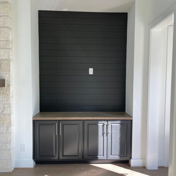 Built-in cabinet with dark doors and a wood countertop set in an alcove with black horizontal shiplap wall, power outlet above—perfect for a kitchen remodel—next to a stone wall and white walls, with sunlight on the wood floor.