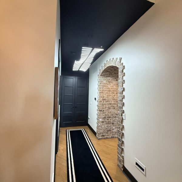 A hallway with white walls, a black ceiling, wood floor, a black area rug with white borders, a brick archway, and black double doors at the end—perfect inspiration for your next remodel, whether it’s the bathroom or kitchen.