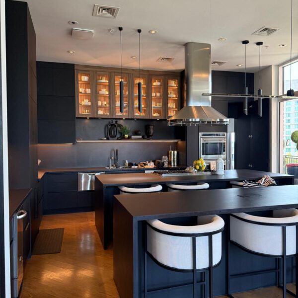 Modern kitchen remodel in The Woodlands features black cabinets, illuminated glass uppers, stainless steel appliances, and a spacious double island with three white cushioned bar stools on wooden floors. Large windows fill the space with natural light.