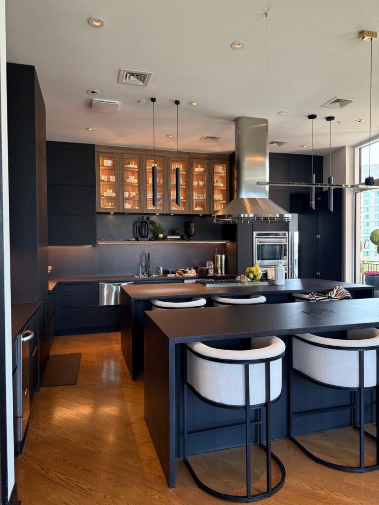 Modern kitchen remodel in The Woodlands features black cabinets, illuminated glass uppers, stainless steel appliances, and a spacious double island with three white cushioned bar stools on wooden floors. Large windows fill the space with natural light.