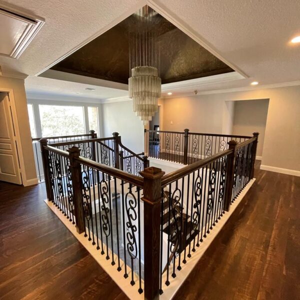 A modern indoor balcony in The Woodlands features dark wooden railings and decorative ironwork, overlooking a lower floor. A large, tiered chandelier hangs from a recessed ceiling, bathing the recent remodel in natural light streaming through spacious windows.