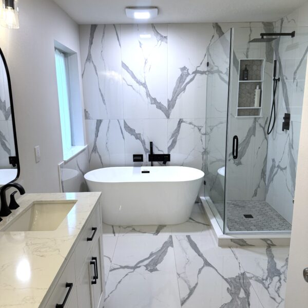 Modern bathroom with white marble floors and walls, a freestanding bathtub, glass-enclosed shower with black fixtures, and a white vanity with a sink and black faucet under a wall mirror.