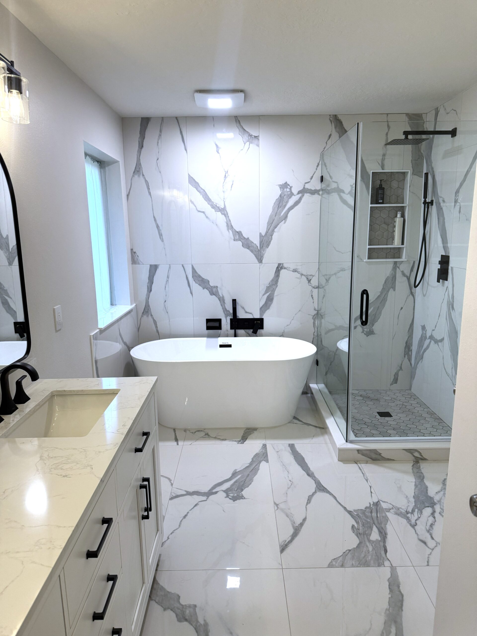 Modern bathroom with white marble floors and walls, a freestanding bathtub, glass-enclosed shower, black fixtures, a large mirror, and a white vanity with a rectangular sink. Natural light comes through a window.