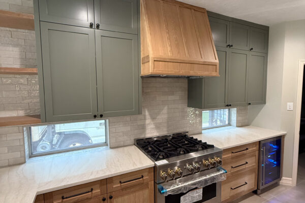 Kitchen with stainless steel gas stove, wood range hood, green upper cabinets, wood lower cabinets, light countertops, gray tile backsplash, and beverage cooler.
