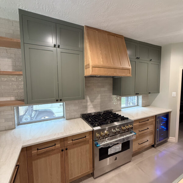 Kitchen with stainless steel gas stove, wood range hood, green upper cabinets, wood lower cabinets, light countertops, gray tile backsplash, and beverage cooler.
