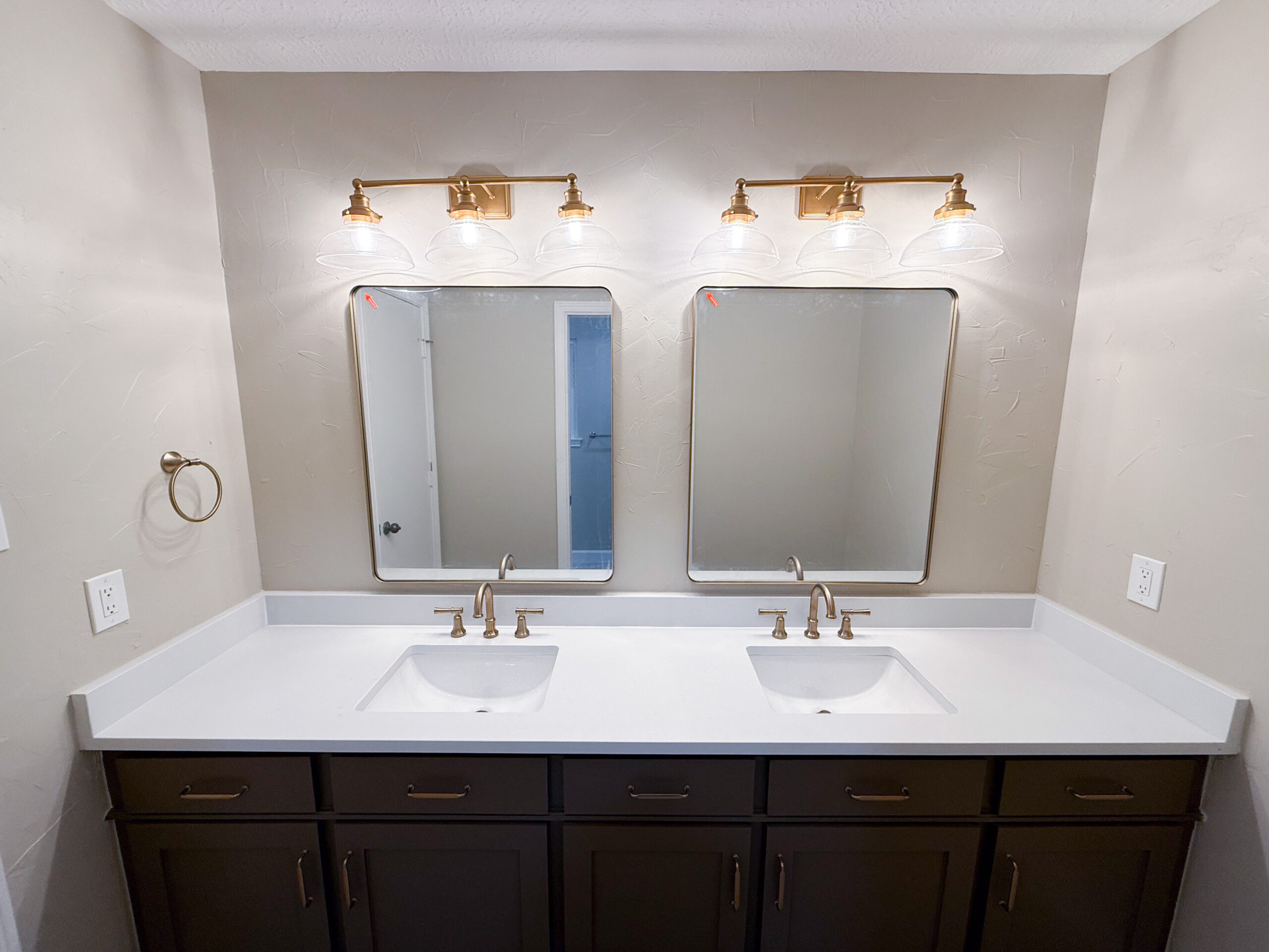 A modern bathroom with two rectangular mirrors, two sinks, and a dark wood vanity. Each sink has a gold faucet, and above each mirror are three-globe gold light fixtures. Remodel done by The Woodlands Premier Remodeling.