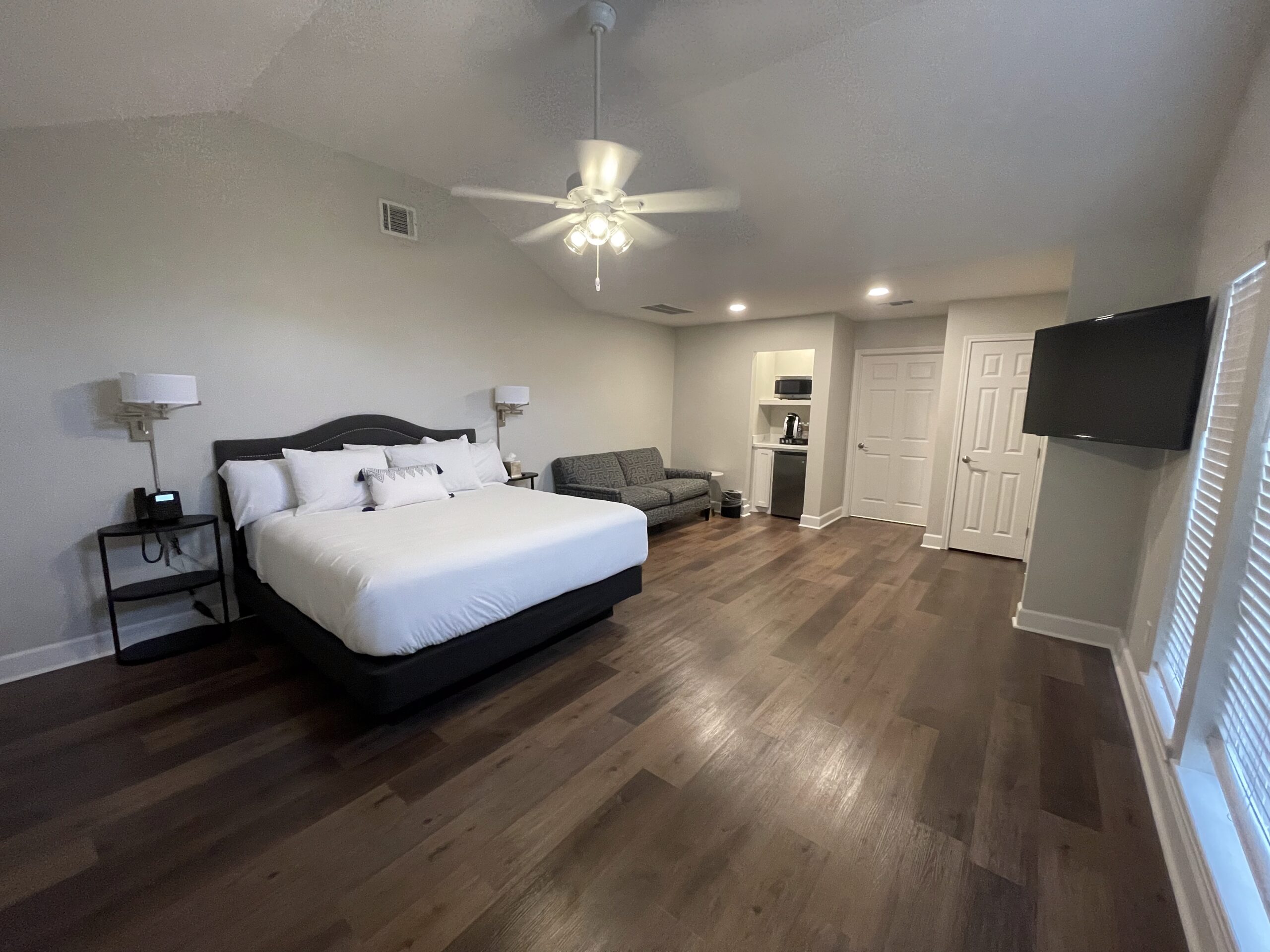 Spacious bedroom remodel with a large bed, gray sofa, wooden floor, mounted TV, ceiling fan, nightstands, and a small kitchenette area near double doors. Large windows let in natural light.