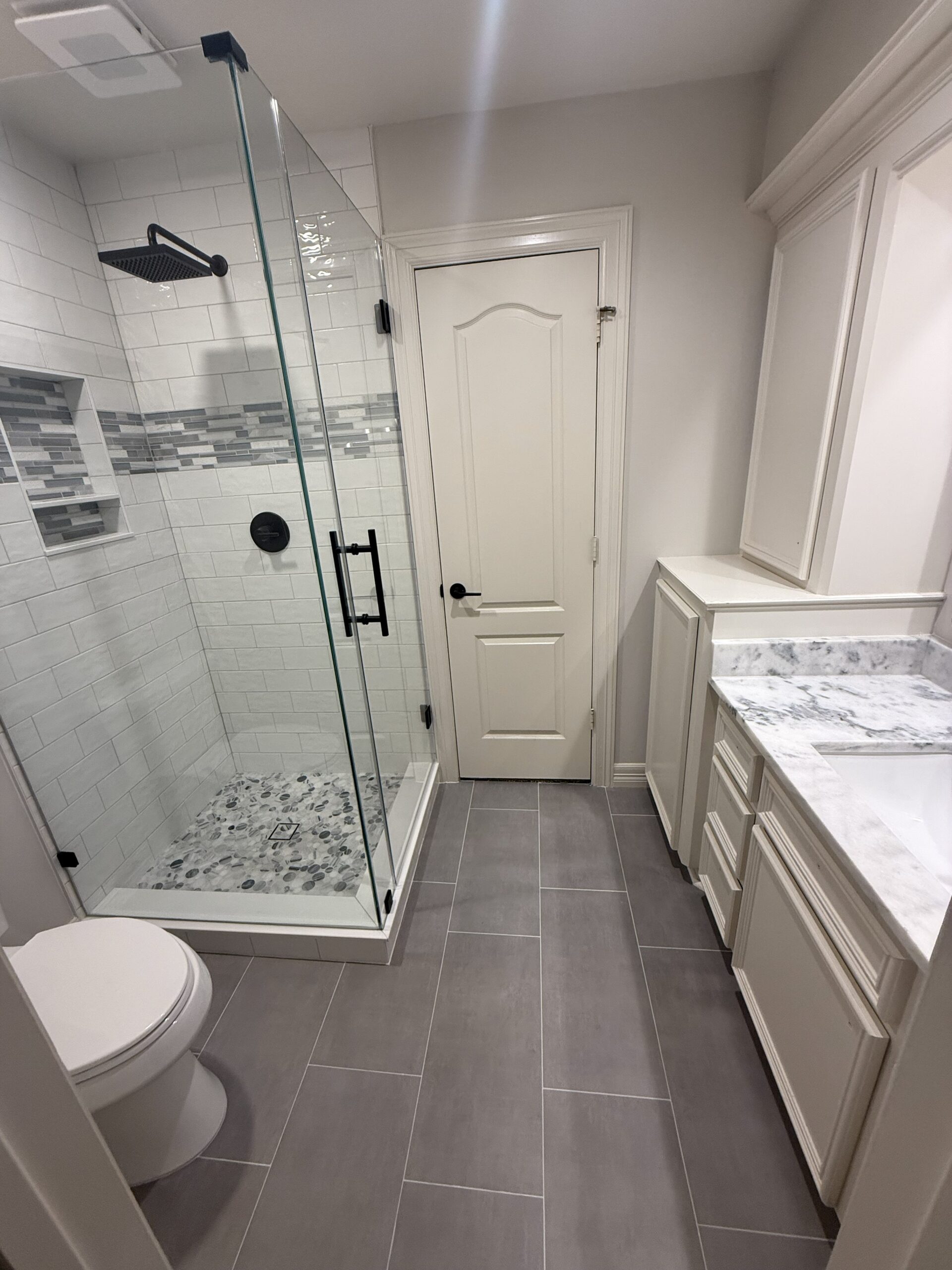 Modern bathroom with gray tile floor, glass-enclosed shower with white subway tiles and gray accents, white toilet, white cabinetry, and marble countertop next to a white door.