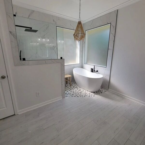 Modern bathroom with light wood floors, freestanding white bathtub, two frosted windows, a small wooden stool, hanging wicker light fixture, and a walk-in shower with marble walls and a partial glass divider. Bathroom remodel done by The Woodlands Premier Remodeling in The Woodlands, TX.