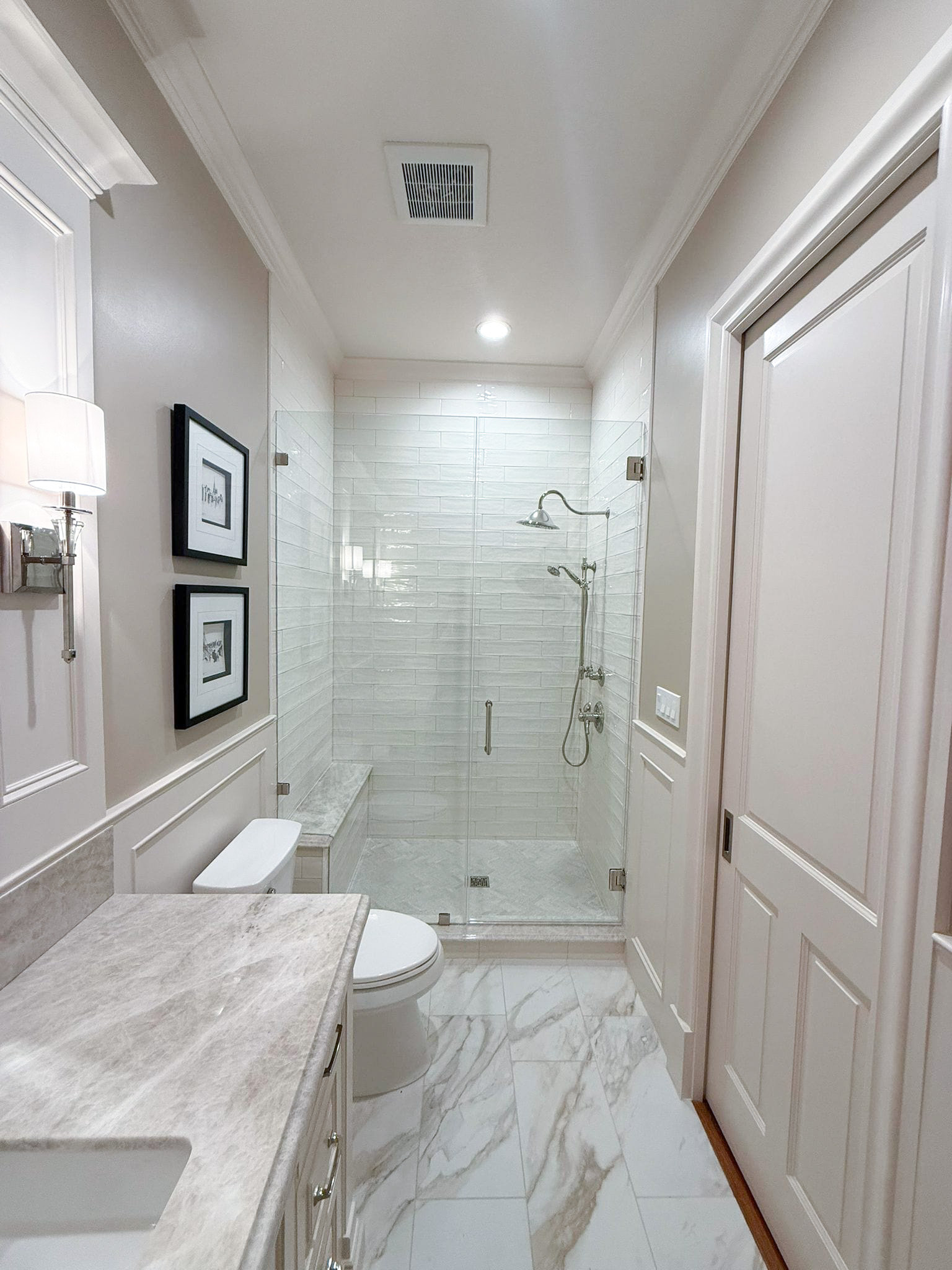 A modern, bright bathroom with marble floors, a glass-enclosed shower, a toilet, and a marble countertop. The walls are light-colored, and framed art decorates one wall above the toilet. Bathroom remodel done by The Woodlands Premier Remodeling in The Woodlands, TX.