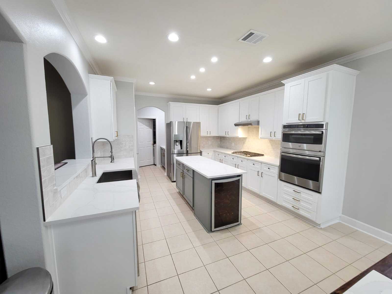 Spacious modern kitchen with white cabinets, stainless steel appliances, a center island with a wine fridge, tiled floor, under-cabinet lighting, and recessed ceiling lights. Kitchen remodel done by The Woodlands Premier Remodeling in The Woodlands, TX.