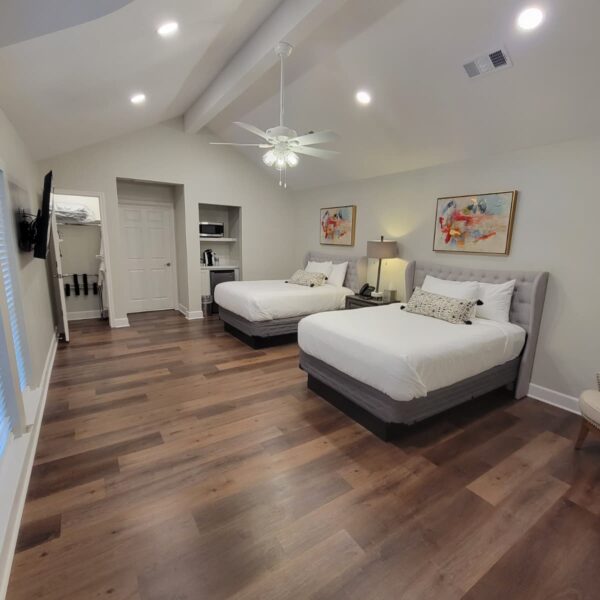 A spacious bedroom with two double beds, white bedding, wood flooring, abstract artwork above each bed, a ceiling fan, recessed lighting, and a door leading to an adjoining room. Remodel done by The Woodlands Premier Remodeling.