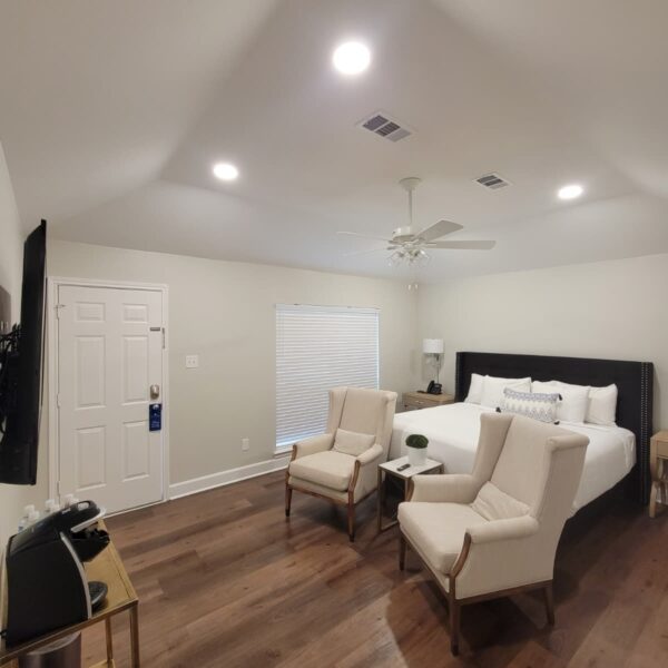 A modern, well-lit bedroom with wood floors, a king-size bed, two beige armchairs with a small table, a TV mounted on the wall, a console with a coffee maker, and a window with closed blinds. Remodel done by The Woodlands Premier Remodeling.