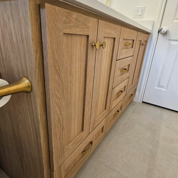 A wooden bathroom vanity with paneled cabinet doors, gold hardware, a white countertop, and a large mirror above, set against a light-colored tiled floor and white walls. Remodel done by The Woodlands Premier Remodeling in The Woodlands, TX.