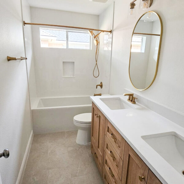 A modern bathroom with a double-sink vanity, gold fixtures, an oval mirror, a toilet, and a bathtub with a gold showerhead under a window. The room features neutral colors and tiled flooring. Remodel done by The Woodlands Premier Remodeling in The Woodlands, TX.