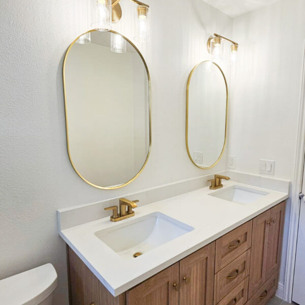 A modern bathroom with a double-sink vanity featuring wood cabinets, gold faucets, two oval mirrors with gold frames, and matching wall-mounted light fixtures above each mirror. A toilet is positioned next to the vanity. Remodel done by The Woodlands Premier Remodeling in The Woodlands, TX.