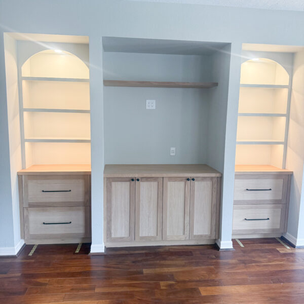 Built-in shelving unit with two light wood bookcases on either side, each with two lower drawers and three shelves, and a central cabinet with double doors below an open shelf, all against a light gray wall with wood flooring. Features under shelf lighting.