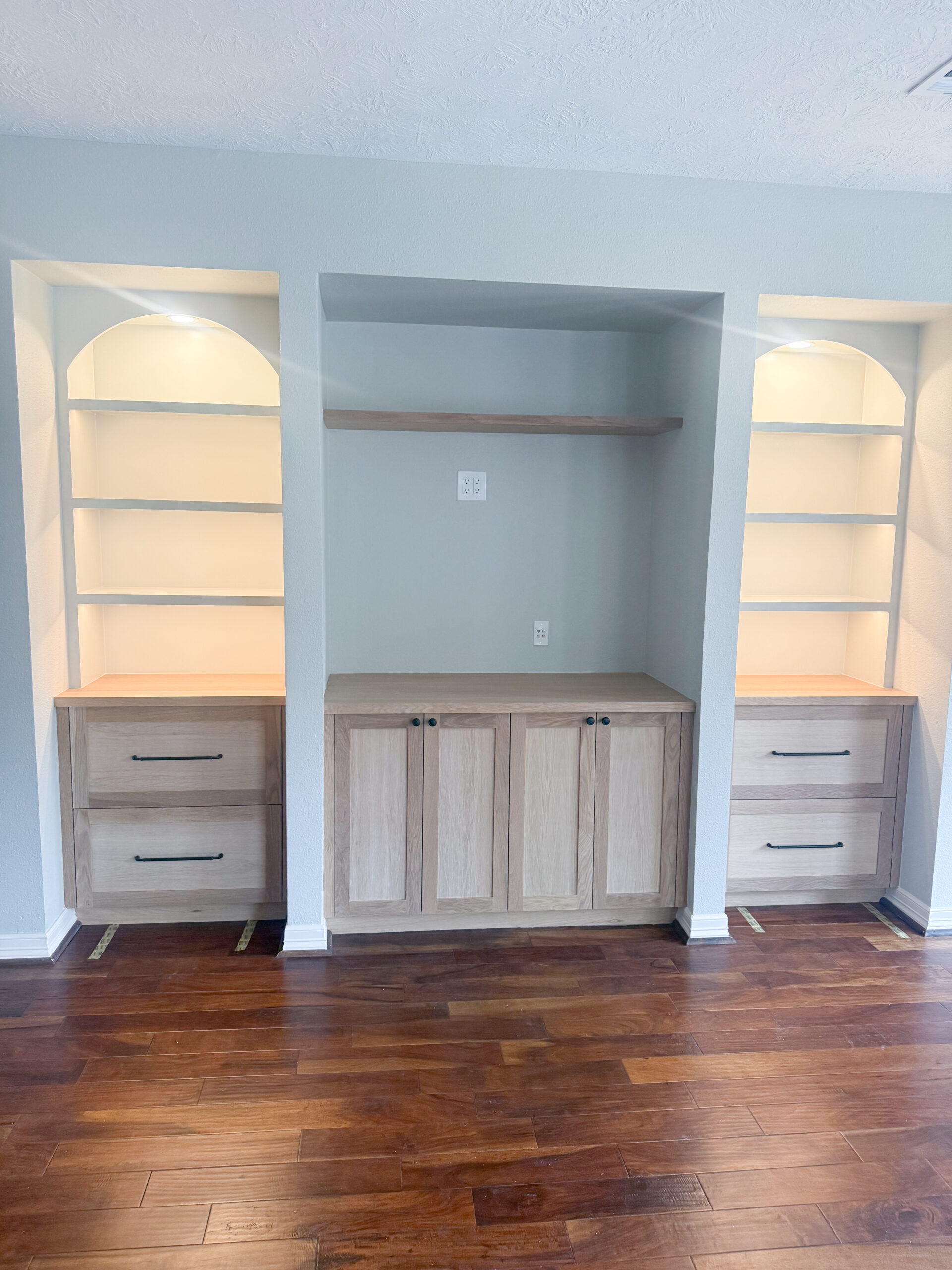 Built-in shelving unit with two light wood bookcases on either side, each with two lower drawers and three shelves, and a central cabinet with double doors below an open shelf, all against a light gray wall with wood flooring. Features under shelf lighting.
