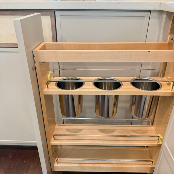 A pull-out kitchen cabinet with wooden shelves holds three stainless steel containers, designed for organizing utensils or kitchen tools. The cabinet is open, revealing its contents and part of the kitchen counter above. Remodel done by The Woodlands Premier Remodel in The Woodlands, TX.