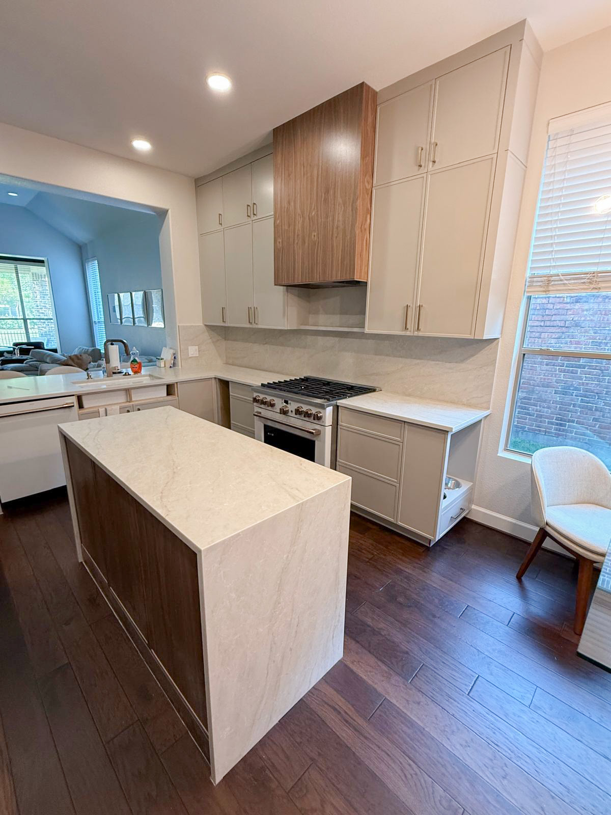 Modern kitchen with light beige cabinets, a marble countertop island with wood accents, a gas stove with oven, and a window with blinds. Wood floors and an open view into a living area are visible.