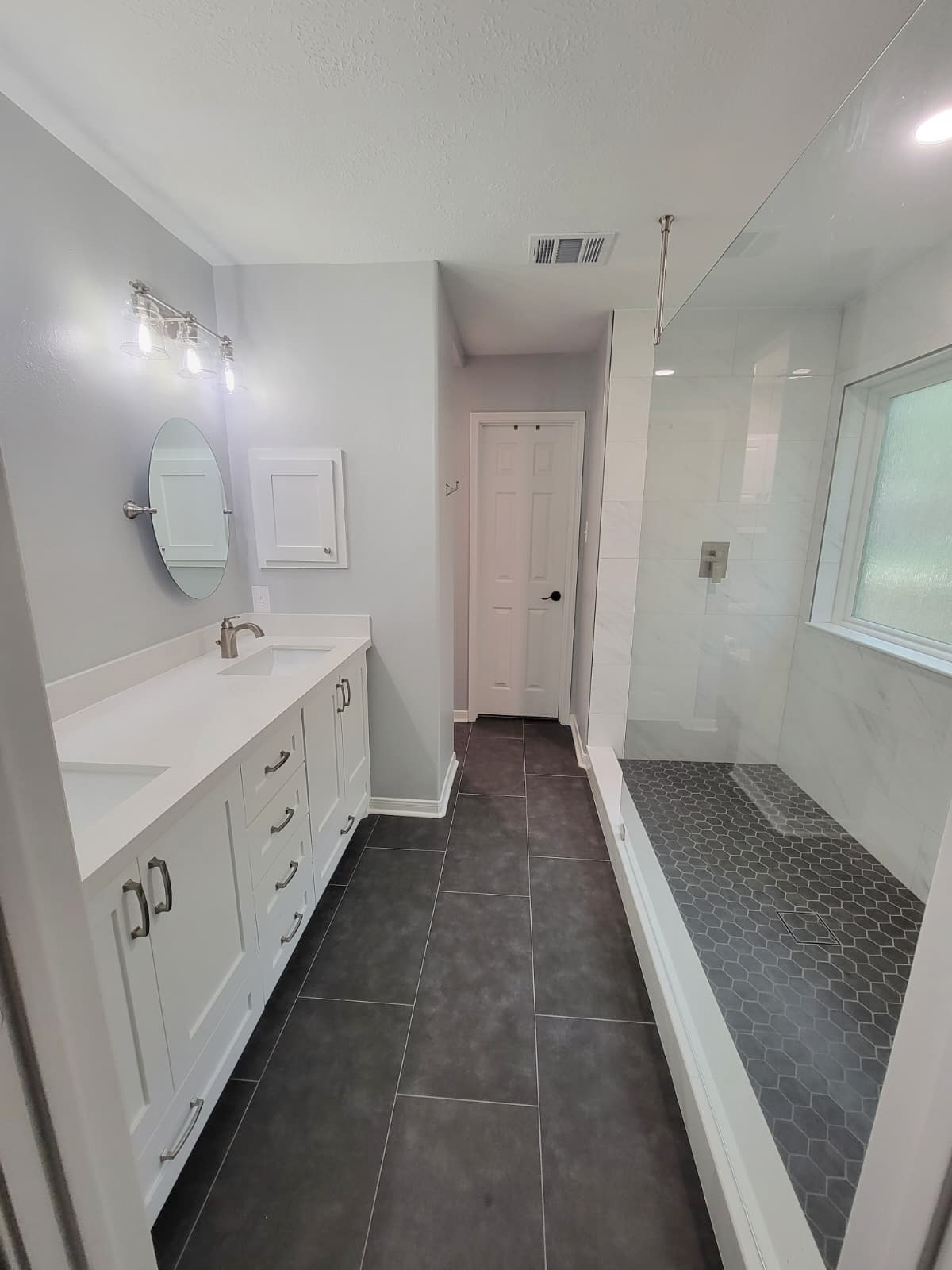 Modern bathroom with white cabinets, dual sinks, a round mirror, gray tile floor, glass shower with dark hexagon tiles, and a frosted window letting in natural light. Remodel done by The Woodlands Premier Remodeling in The Woodlands, TX.