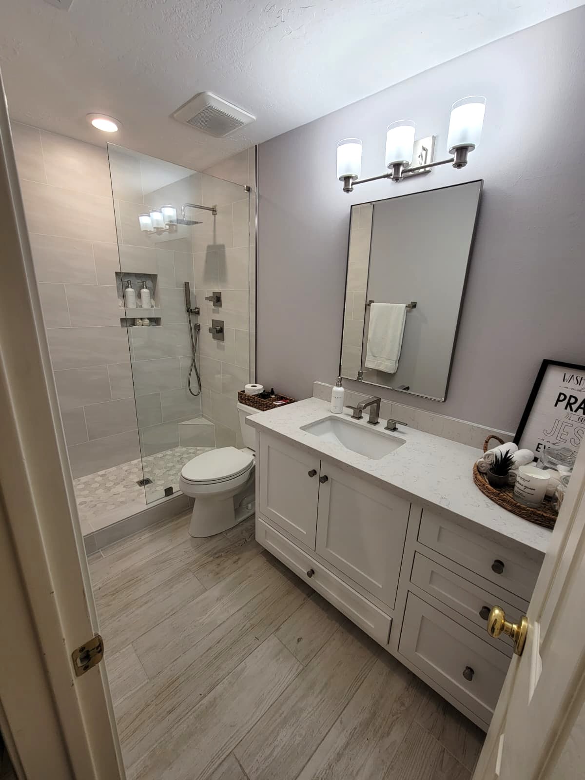 Modern bathroom with light gray walls, a glass-enclosed shower, white vanity with a sink, large mirror, three-light fixture above, toilet, and decorative tray with various items on the countertop. Light wood-look floor tiles. Bathroom remodel done by The Woodlands Premier Remodeling in The Woodlands, TX.