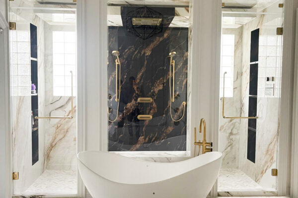 A modern bathroom remodeling features a white freestanding tub, gold fixtures, two glass-enclosed showers on either side, marble walls and floors, and a geometric black chandelier overhead.