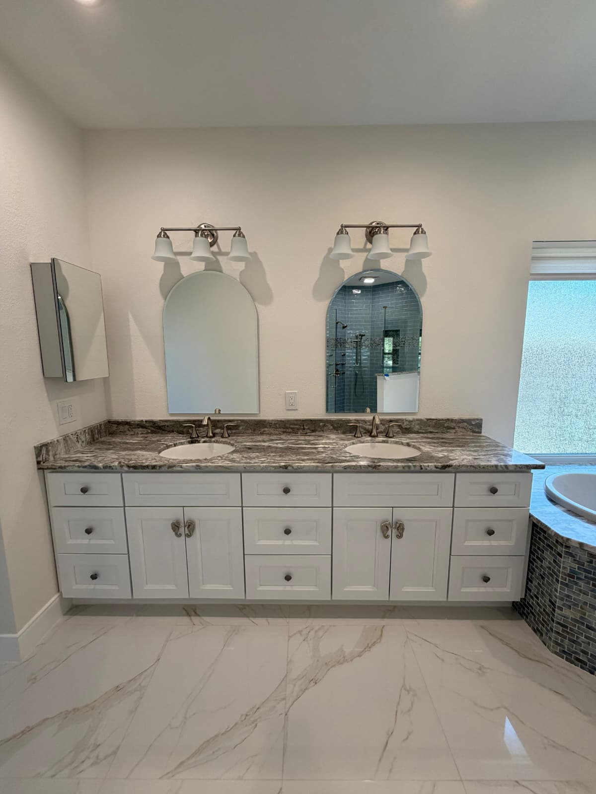 Modern bathroom with white double vanity, marble countertop, two arched mirrors, and two light fixtures above. There’s tiled flooring, built-in cabinets, and a partial view of a bathtub with mosaic tile on the right. Bathroom remodel by The Woodlands Premier Remodeling in The Woodlands, TX.