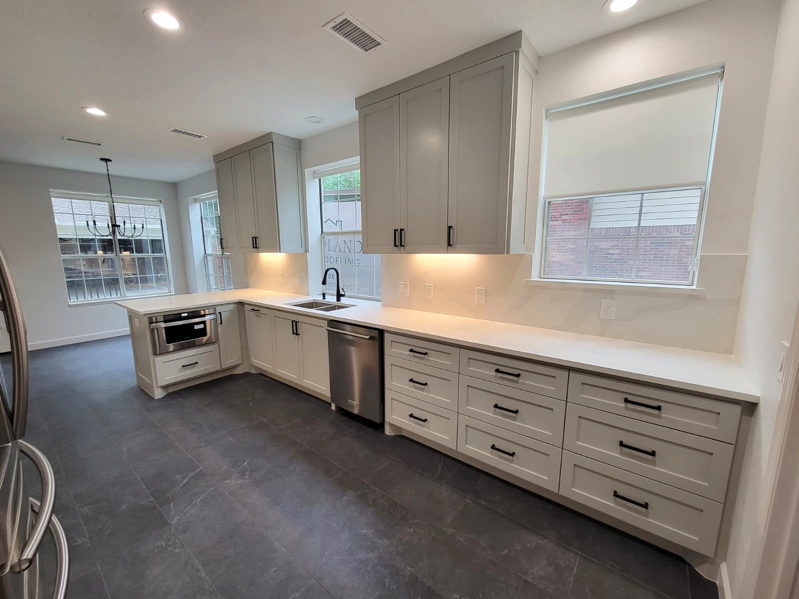Modern kitchen with white cabinets, black handles, white countertops, stainless steel appliances, large windows, and dark tile flooring. The room has bright lighting and a clean, minimalist design. Remodel done by The Woodlands Premier Remodeling in The Woodlands TX.