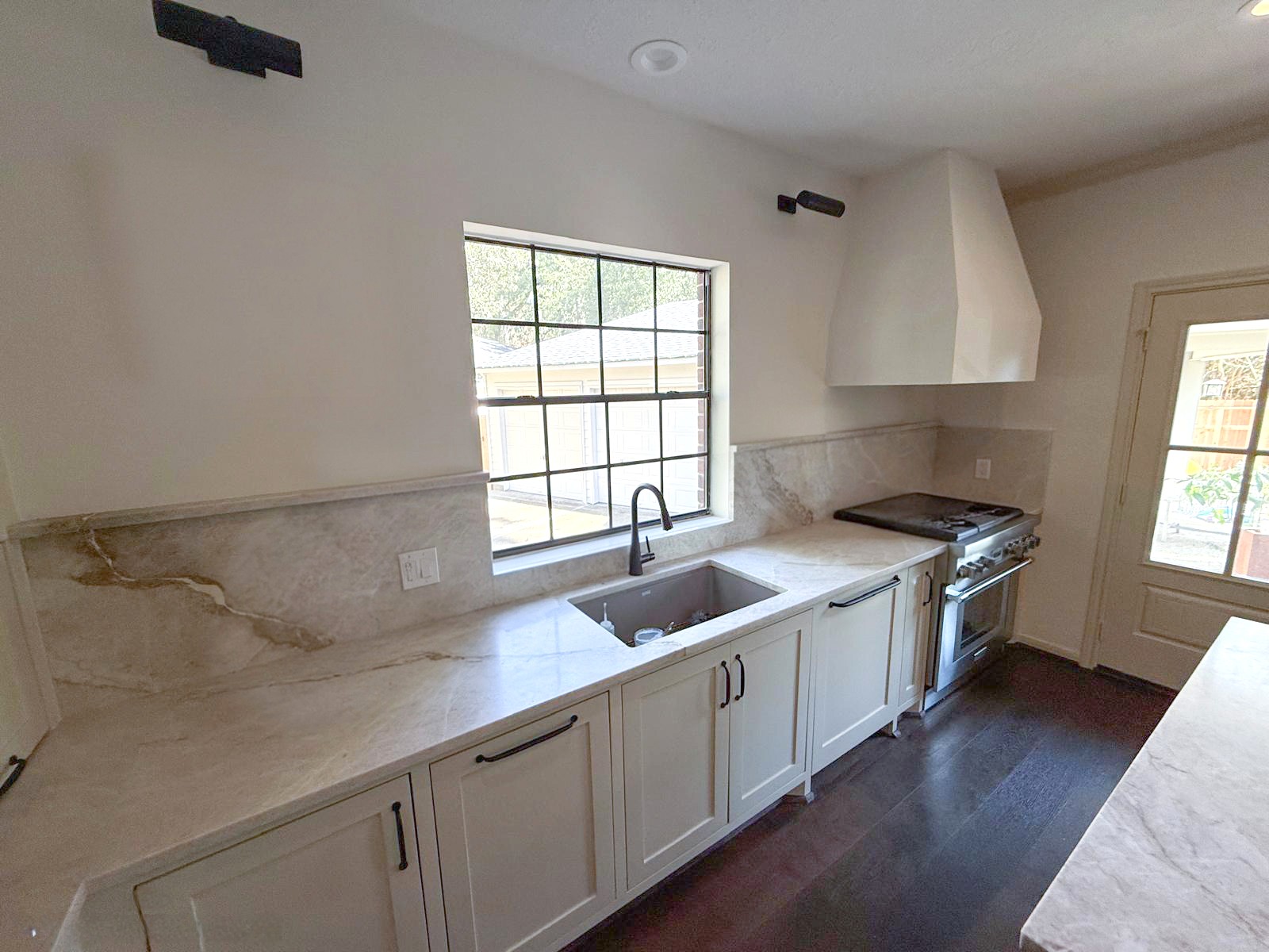 Modern kitchen with white cabinets, marble countertops, a stainless steel sink under a window, and a gas stove with a white range hood; sunlight streams in through the window and door. Remodel done by The Woodlands Premier Remodeling.