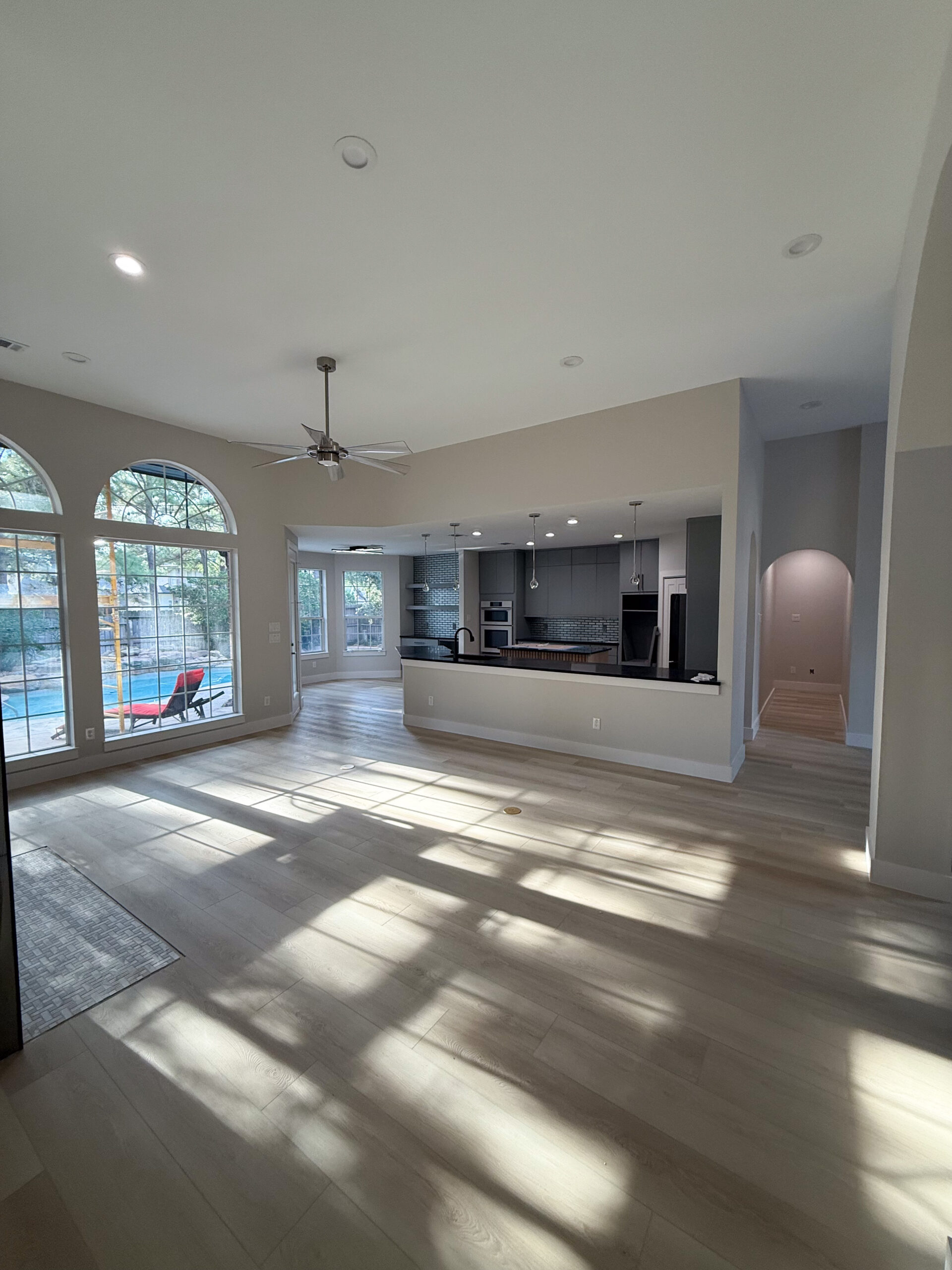 Spacious modern living area with light wood floors, large arched windows overlooking a pool, open kitchen with island, and hallway entrance. Sunlight streams in, creating bright, airy atmosphere.