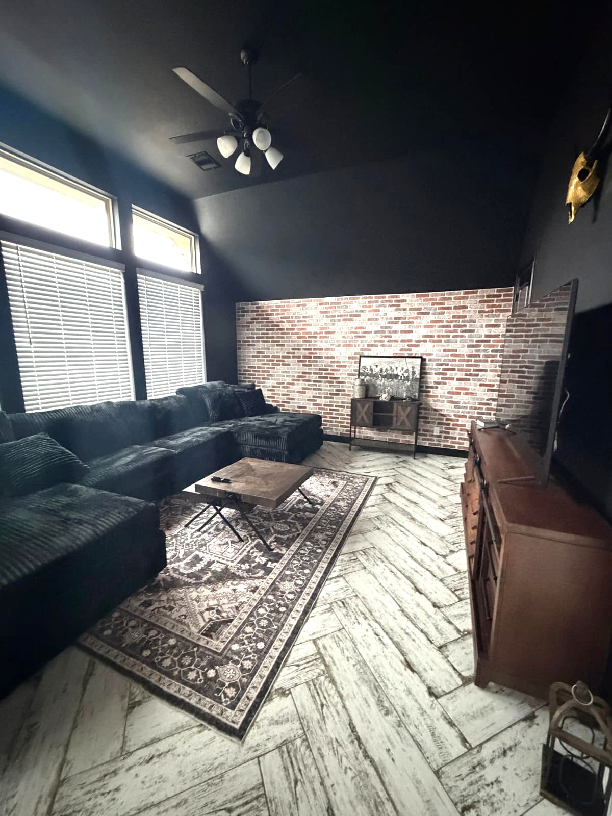 A cozy home theater remodel with dark walls, a large black sectional sofa, a patterned rug, a wooden coffee table, a brick accent wall, and a TV on a wooden stand. Large windows let in natural light.