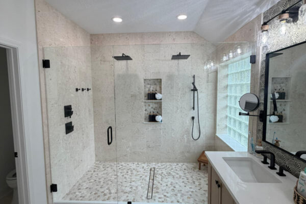Modern bathroom remodel with a spacious glass-enclosed shower featuring two showerheads, built-in shelves with toiletries, a wood bench, double-sink vanity with a large mirror, and a black and white patterned rug on the floor.