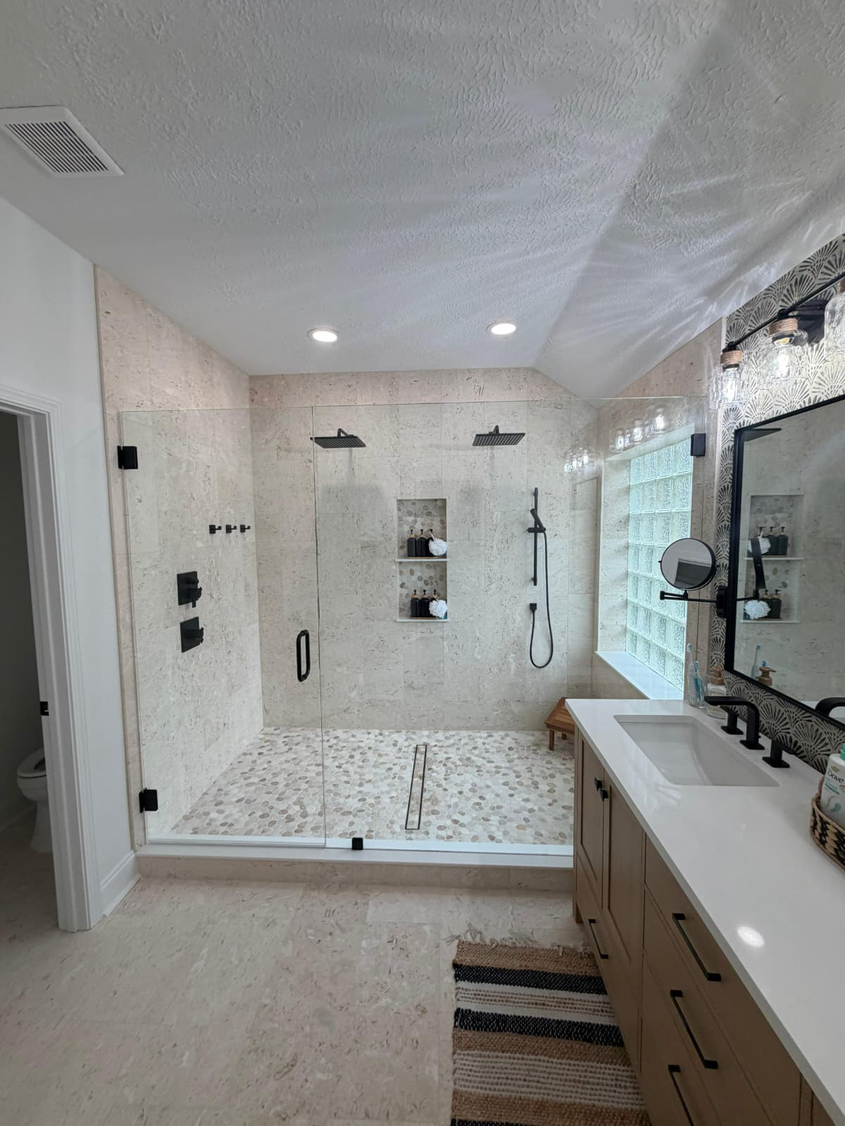 Modern bathroom remodel with a spacious glass-enclosed shower featuring two showerheads, built-in shelves with toiletries, a wood bench, double-sink vanity with a large mirror, and a black and white patterned rug on the floor.