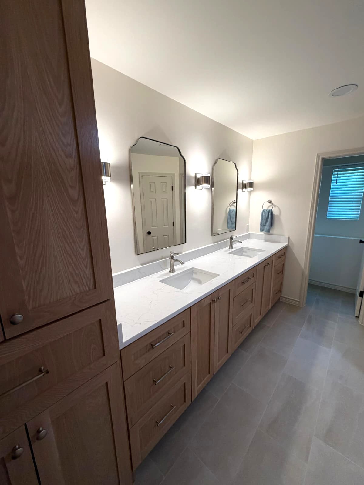 A modern bathroom with light wood cabinets, a white double-sink vanity, two rectangular mirrors with curved tops, wall-mounted lights, and a small window in the background. The floor is covered with large gray tiles.