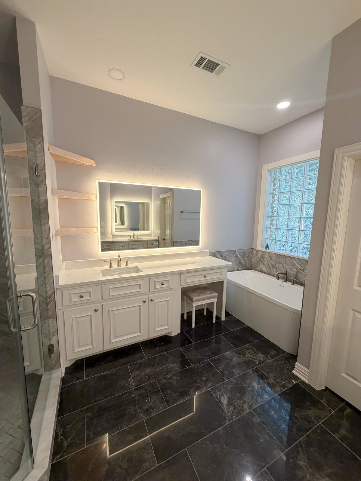 Modern bathroom with a large backlit mirror, white vanity, black tile floor, soaking tub by a glass block window, and a glass-enclosed shower on the left.