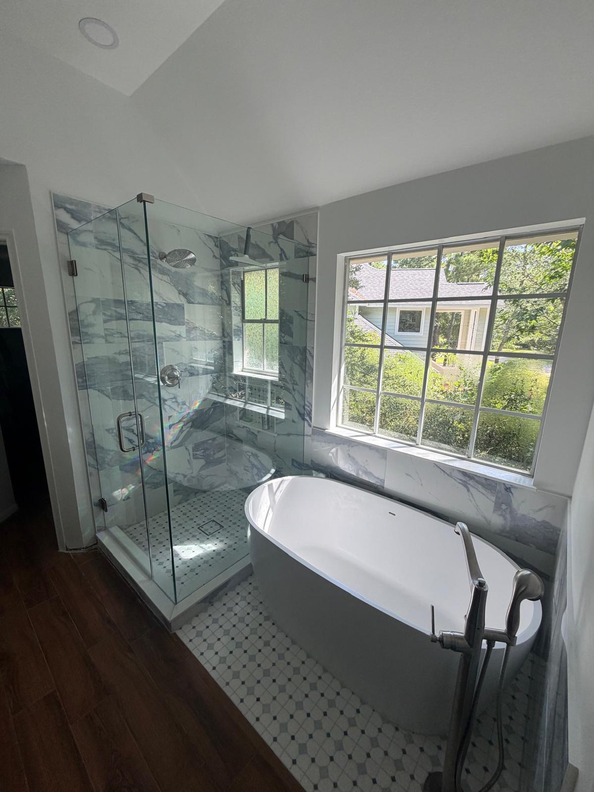 Modern bathroom remodel with a glass-enclosed shower, white freestanding tub, marble-patterned walls, wood floor, and a large window letting in natural light with greenery outside.
