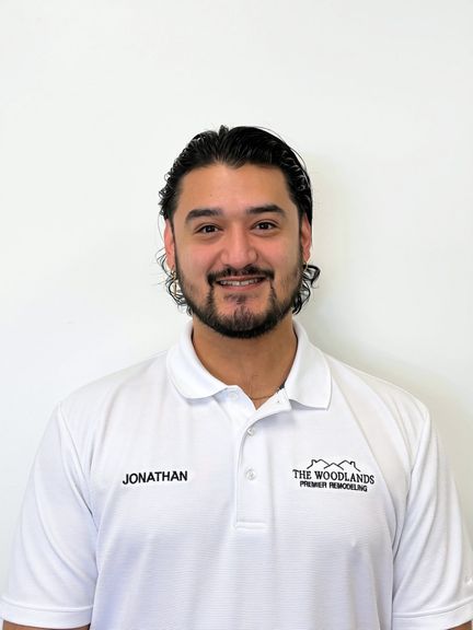 A man with dark hair and a beard smiles at the camera. He is wearing a white polo shirt with JONATHAN and THE WOODLANDS PREMIER REMODELING logos on it, standing against a plain white background, reflecting the dedication and family values that define his work.
