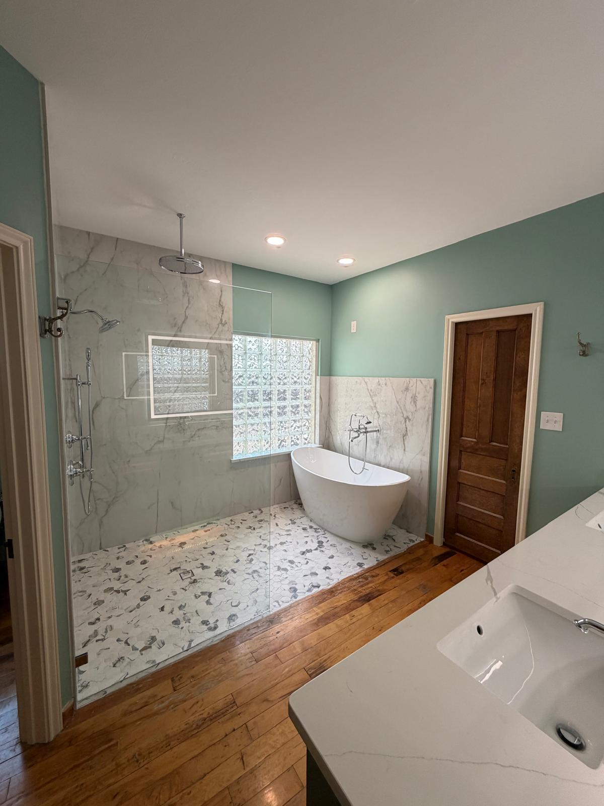 Modern bathroom with a glass walk-in shower, rainfall showerhead, freestanding tub, wooden door, dual sinks, marble accents, wooden floor, and a large glass block window letting in natural light.