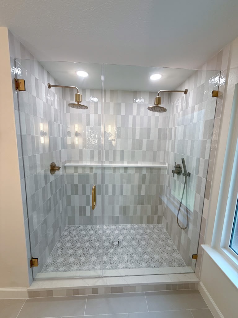 Modern double shower with two rainfall showerheads, patterned tile floor, vertical rectangular wall tiles, glass doors, gold hardware, and a window on the right side. The space is well-lit with ceiling lights.